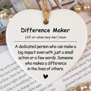 Könnte beinhalten: Ein weißes herzförmiges Keramikornament mit dem Text "Difference Maker" und einer Definition. Das Ornament hängt an einer Juteschnur und ist von Kunstperlen und Blumen umgeben. Ein nachdenkliches Geschenk.
