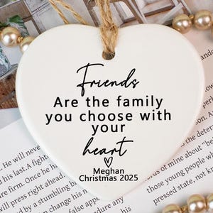 May include: A white heart-shaped ornament with a jute string for hanging. The ornament features the text "Friends Are the family you choose with your heart" and "Meghan Christmas 2025".