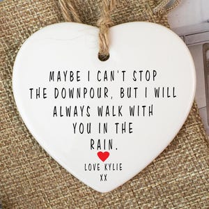 May include: A white heart-shaped ornament with a jute string. The ornament features the text "MAYBE I CAN'T STOP THE DOWNPOUR, BUT I WILL ALWAYS WALK WITH YOU IN THE RAIN. LOVE KYLIE XX" in black. It is displayed on a burlap bag.