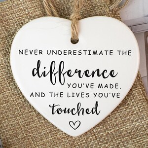 May include: A white heart-shaped ornament with a jute string. The ornament features the text "NEVER UNDERESTIMATE THE difference YOU'VE MADE, AND THE LIVES YOU'VE touched" in black script. It is placed on a burlap bag.