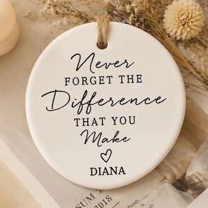 Könnte beinhalten: Ein rundes, weißes Keramikornament mit einer Juteschnur zum Aufhängen. Der Text lautet: Never FORGET THE Difference THAT YOU Make, gefolgt von einem kleinen Herzsymbol und dem Namen DIANA in schwarzer Schrift.