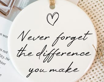 Puede incluir: Un adorno redondo de cerámica blanca con una cuerda de yute para colgar. El adorno presenta el texto "Never forget the difference you make" en escritura negra, con un pequeño corazón encima. El fondo incluye flores y una cinta blanca.