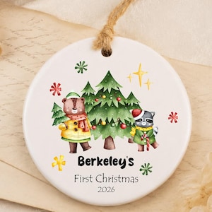 Könnte beinhalten: Ein rundes, weißes Keramikornament mit einer Aquarellillustration eines Bären und eines Waschbären neben einem Weihnachtsbaum. Der Text lautet Berkeley's First Christmas 2026. Das Ornament hängt an einer braunen Kordelschlaufe.