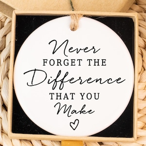 Könnte beinhalten: Ein weißes, rundes Keramikornament mit dem Text "Never Forget The Difference That You Make" in schwarzer Schrift. Das Ornament befindet sich in einer kleinen Schachtel mit dem Aufkleber "Hand made". Der Hintergrund umfasst eine gewebte Matte und florale Akzente.
