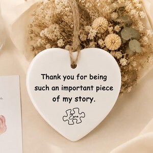 May include: A white heart-shaped ceramic ornament hanging from a jute twine string. The ornament features the black printed text, Thank you for being such an important piece of my story, and a puzzle piece icon with the name Zora written inside.