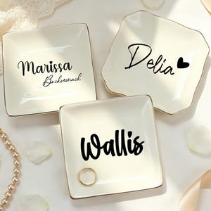 Könnte beinhalten: Drei quadratische, cremefarbene Keramikschalen mit goldfarbenen Rändern. Jede Schale trägt schwarzen Text: eine mit Marissa Bridesmaid, eine mit Delia und einem Herzsymbol sowie eine mit Wallis. Ein goldener Ring liegt auf der Wallis-Schale.