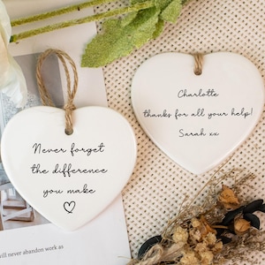 May include: Two white ceramic heart decorations with jute twine loops. One says "Never forget the difference you make" with a heart symbol. The other says "Charlotte thanks for all your help! Sarah xx". A small dried flower bouquet is at the bottom.