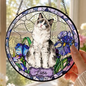 Könnte beinhalten: Ein runder Buntglas-Sonnenfänger mit einer weiß-grauen Katze mit blauen Augen, die zwischen violetten und blauen Schwertlilien sitzt. Das Stück hat einen schwarzen Metallrahmen, eine Aufhängungskette und ein violettes Namensschild mit dem Text Luna.