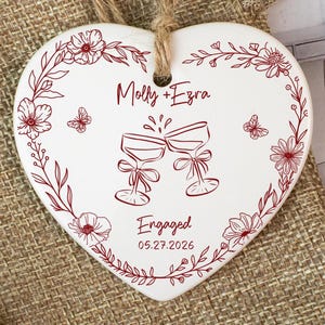 Könnte beinhalten: Ein weißes, herzförmiges Keramikornament mit rotem Blumenrand, zwei anstoßenden Weingläsern und dem Text "Molly + Ezra" sowie "Engaged 05.27.2026". Das Ornament hängt an einer Juteschnur über einem kleinen Beutel aus Jute.