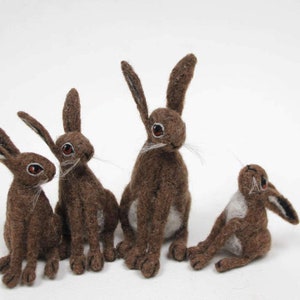 Puede incluir: Cuatro figuritas de liebre de fieltro marrón, cada una con una pose diferente. Las liebres tienen pelo blanco en el vientre y orejas largas.