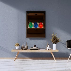 Puede incluir: Un expositor enmarcado en madera con un cofre abierto que contiene tres gemas brillantes. Las gemas son verdes con un rayo, azules y rojas. El expositor está en una pared azul sobre una mesa de madera con objetos decorativos.
