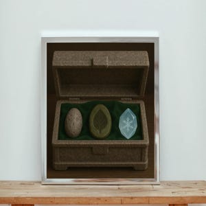 May include: A framed image showing a brown box with an open lid, displaying three items on a green cloth. These include a speckled egg, a leaf-shaped object, and a blue crystal. The box is placed on a wooden surface.