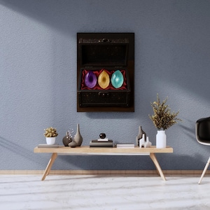 Op de afbeelding: Een zwarte houten kist met een open deksel, met drie kleurrijke, edelsteenachtige stenen: paars, goud en blauwgroen. De kist is aan een blauwe muur bevestigd. De edelstenen liggen op een rode fluwelen voering.
