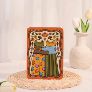 May include: An orange rectangular art print featuring two hugging frogs. One frog wears a brown sweater and polka-dot pants, the other a blue sweater and striped pants. The border includes pumpkins, mushrooms, and leaves.