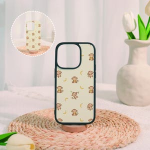 May include: A cream-coloured phone case featuring a repeating pattern of small brown cartoon monkeys and yellow bananas. The case has a black border around the camera cutout and the outer edge, with a circular inset showing the same design.