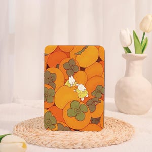 Può includere: Un oggetto decorativo rettangolare con una vivace illustrazione di cachi nei toni dell'arancione e del verde. Due personaggi dei cartoni animati in giallo e bianco interagiscono. L'oggetto è posto su un tappetino intrecciato. Probabilmente è una custodia per tablet.
