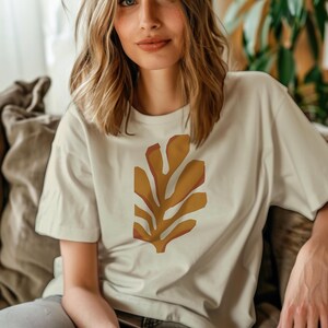 May include: Cream-colored t-shirt featuring a large, abstract leaf design in shades of brown and orange. The short-sleeved shirt is made of a soft material and is suitable for casual wear. The design is centered on the front of the shirt.