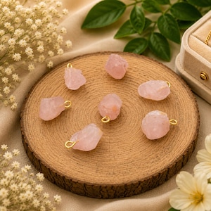 May include: A collection of six raw rose quartz pendants, each featuring an irregular, translucent pale pink stone attached to a small gold-toned metal jump ring. The stones are arranged in a circular pattern on a round, textured wooden slice.