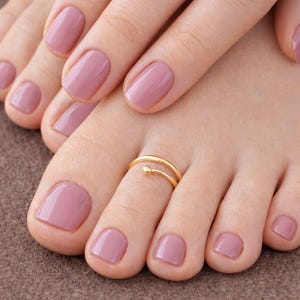 Könnte beinhalten: Nahaufnahme eines Fußes und einer Hand mit mauvefarbenem Nagellack. Ein goldener Zehenring mit kleinem Kugelakzent wird am großen Zeh getragen. Die Fingernägel sind ebenfalls mauvefarben lackiert.