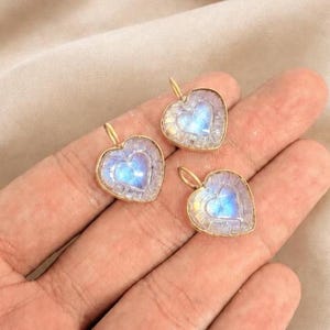May include: Three heart-shaped pendants featuring iridescent moonstone centres with a blue glow, encased in gold-toned metal frames with a textured, segmented border design, each equipped with a small gold-toned loop for attachment.