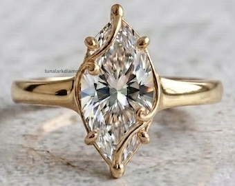 May include: A gold ring featuring a marquise-cut clear gemstone set in a delicate metal frame with curved prongs. The band is smooth and polished gold. The text lunalarkdiamonds is visible in small black letters near the center of the ring.