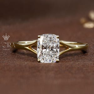 Op de afbeelding: Een gouden verlovingsring met een grote, rechthoekig geslepen diamant. De ring heeft een split-schacht ontwerp, waarbij de band zich splitst in twee delicate tanden die de diamant vasthouden. De ring is geplaatst tegen een bruine achtergrond.