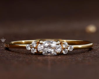 May include: A delicate gold ring featuring a central oval-cut diamond, flanked by smaller, round accent stones. The ring's band is slender, and the overall design is elegant and minimalist. The ring is set against a dark brown background.