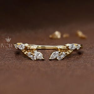 Könnte beinhalten: Ein goldfarbener Ring mit einem einzigartigen Design. Der Ring verfügt über eine Reihe kleiner, klarer Edelsteine, die in einem blattartigen Muster angeordnet sind. Der Ring ist vor einem braunen Hintergrund platziert, der seine komplizierten Details hervorhebt. Der Ring ist ein Schmuckstück.