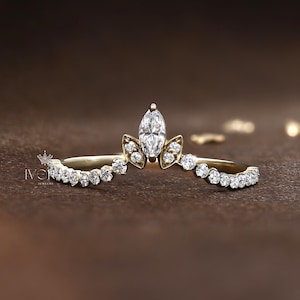 May include: A gold ring set with a marquise-cut diamond and smaller round diamonds. The ring has a crown-like design with a central marquise diamond and a curved band. The ring is set on a brown surface.