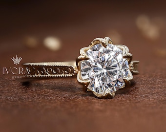 Op de afbeelding: Een gouden verlovingsring met een grote, heldere, rond geslepen diamant in een bloemvormige zetting. De band heeft een delicaat, gegraveerd ontwerp. De ring wordt gepresenteerd op een bruin oppervlak. De tekst "IVORA" is zichtbaar.