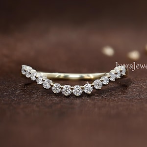 May include: A curved, gold-toned ring set with a series of small, round, clear gemstones. The ring's design features a gentle arc, with the gemstones arranged along the curve. The background is a warm, brown tone.