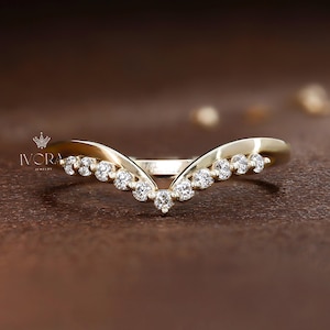 Puede incluir: Un anillo dorado curvado con una fila de pequeñas piedras preciosas transparentes. El anillo tiene un diseño en forma de V, con las piedras preciosas engastadas a lo largo de la curva. El anillo está sobre una superficie marrón.