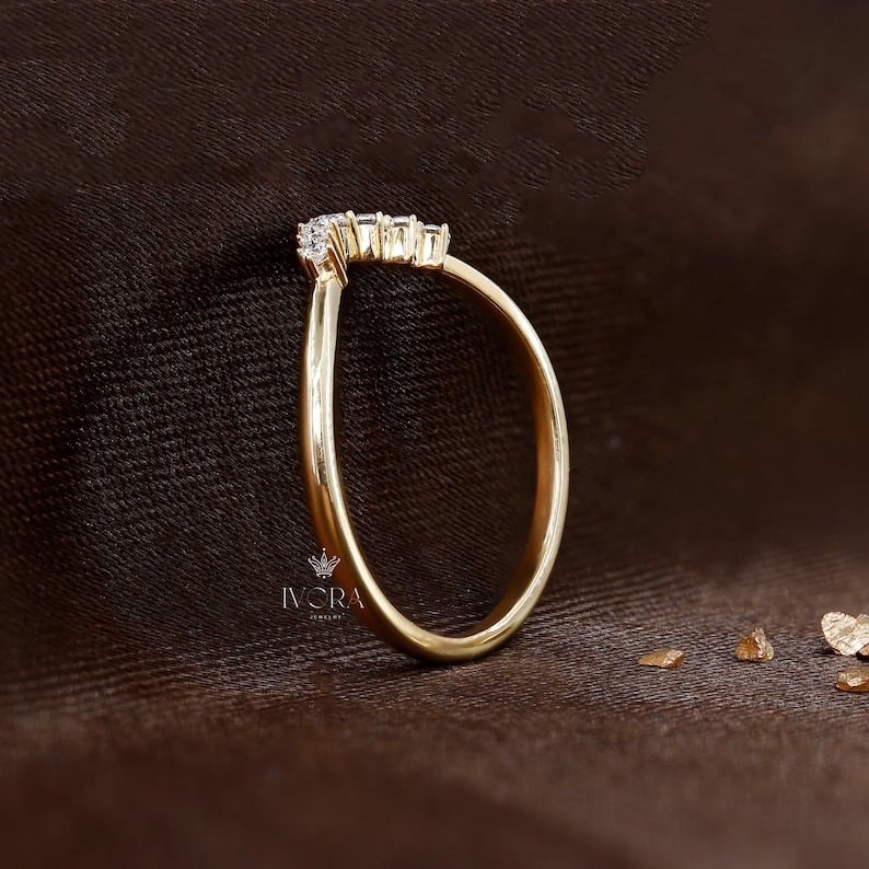 Puede incluir: Un anillo de oro con un dise&ntilde;o curvo, engastado con peque&ntilde;as piedras preciosas transparentes. El anillo se muestra sobre una superficie marr&oacute;n, con el logotipo de joyer&iacute;a IVORA visible. La banda del anillo es lisa y pulida.