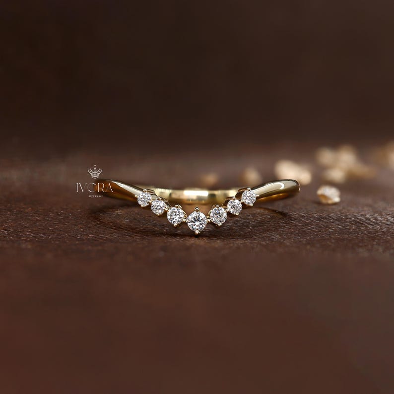 Puede incluir: Un anillo de oro curvado con siete diamantes redondos y transparentes. El anillo tiene un acabado pulido y se encuentra sobre un fondo marr&oacute;n. Es una joya delicada.