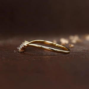 Puede incluir: Un anillo dorado con un dise&ntilde;o curvo. El anillo presenta un grupo de peque&ntilde;as piedras preciosas transparentes en la parte superior, a&ntilde;adiendo brillo. El anillo se encuentra sobre un fondo marr&oacute;n borroso, resaltando la elegante forma de la joya.