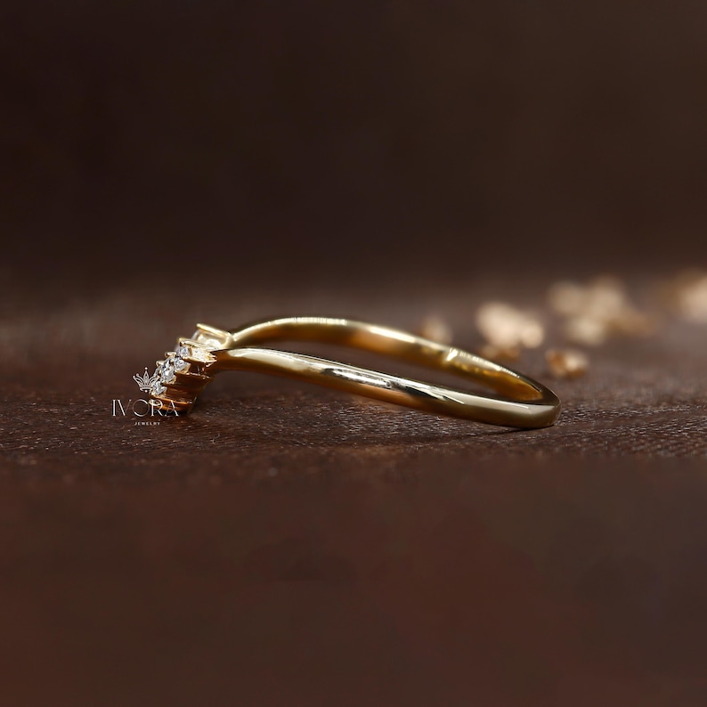 Puede incluir: Un anillo dorado con un dise&ntilde;o curvo. El anillo presenta un grupo de peque&ntilde;as piedras preciosas transparentes en la parte superior, a&ntilde;adiendo brillo. El anillo se encuentra sobre un fondo marr&oacute;n borroso, resaltando la elegante forma de la joya.