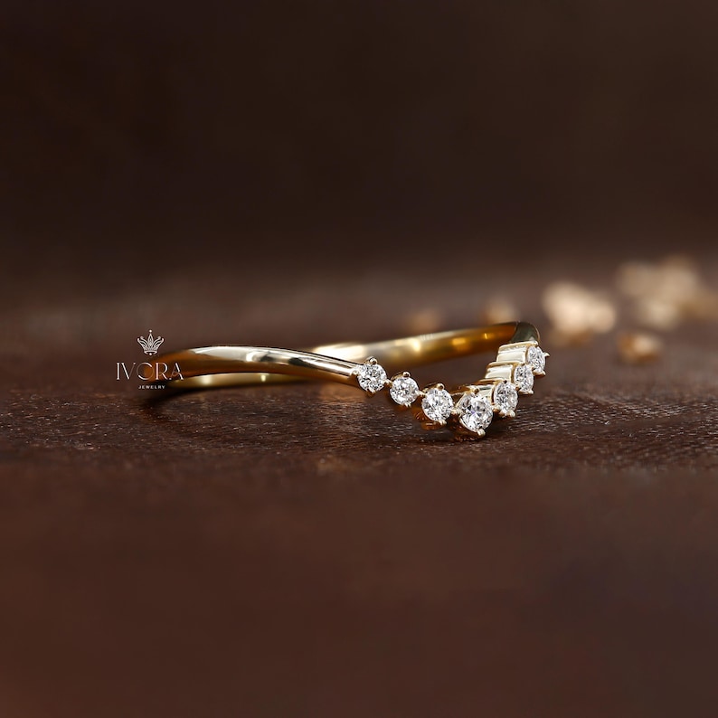 Puede incluir: Un anillo de oro curvado con una fila de peque&ntilde;as piedras preciosas redondas y claras. El anillo tiene un dise&ntilde;o delicado y elegante. El anillo est&aacute; sobre un fondo marr&oacute;n oscuro. La marca "IVORA" es visible.
