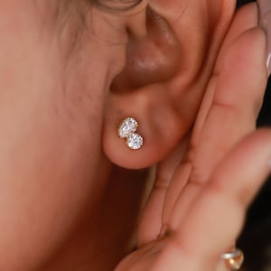 Pu&ograve; includere: Primo piano di un set di orecchini a bottone in oro. L'orecchino presenta una pietra trasparente a forma di pera e una pietra trasparente rotonda. L'orecchino &egrave; incastonato in una montatura in metallo dorato. L'orecchino &egrave; sull'orecchio di una persona.