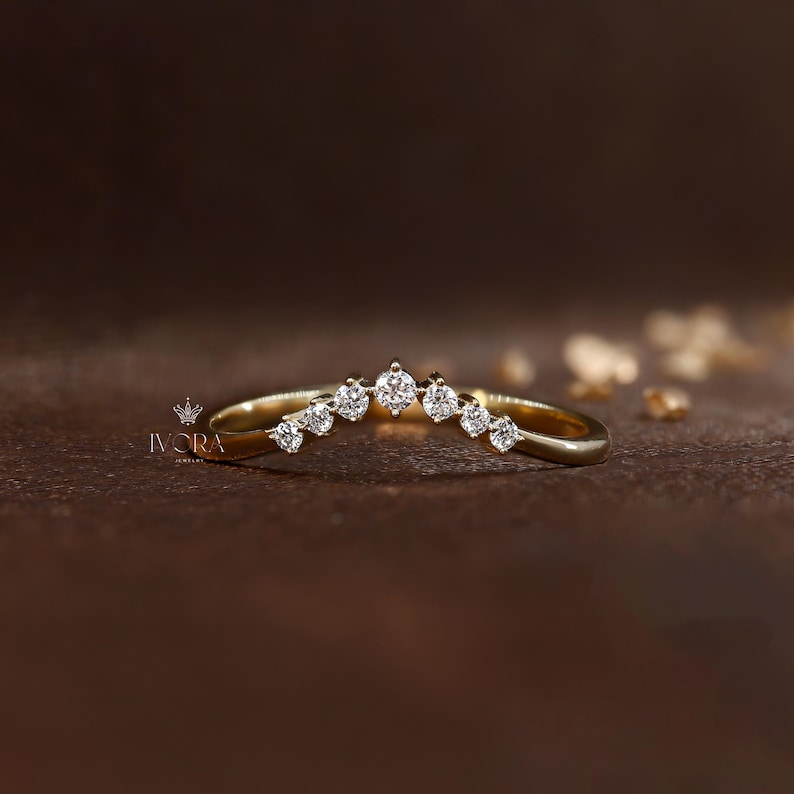 Puede incluir: Un anillo de oro curvado con siete piedras preciosas transparentes. El anillo tiene un dise&ntilde;o delicado, con las piedras preciosas dispuestas en una fila a lo largo de la curva superior. El anillo se muestra sobre una superficie marr&oacute;n.
