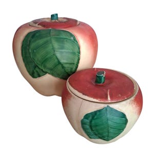 May include: Two ceramic apple-shaped containers with lids. The apples are painted with a red top, a green stem, and a green leaf. The body of the apples is off-white with a pinkish-red blush. One container is larger than the other.
