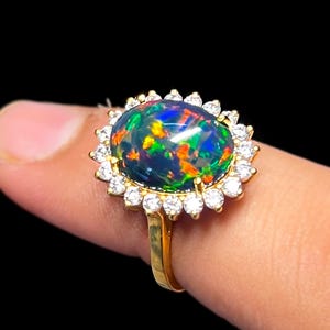 May include: A gold ring featuring a large, oval black opal gemstone with vibrant flashes of color. The opal is surrounded by a halo of small, clear gemstones. The ring's band is a simple, polished gold.