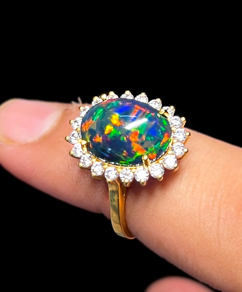 May include: A gold ring featuring a large, oval black opal gemstone with vibrant flashes of color. The opal is surrounded by a halo of small, clear gemstones. The ring's band is a simple, polished gold.