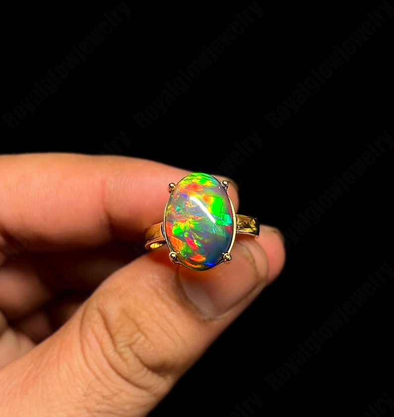 May include: A gold ring featuring a large, oval-shaped opal gemstone. The opal displays a vibrant play of color, with flashes of green, red, and yellow. The ring's band is a simple, polished gold design.