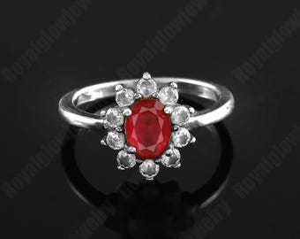 Op de afbeelding: Zilveren ring met een grote ovale robijn in het midden, omringd door kleinere, heldere edelstenen in een bloemvormig ontwerp. De ring heeft een eenvoudige, gepolijste zilveren band. De ring staat tegen een zwarte achtergrond.