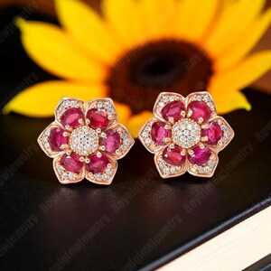 May include: A pair of flower-shaped stud earrings. Each earring features a cluster of oval-cut, deep red gemstones, surrounded by smaller, clear stones. The center of each flower is a cluster of small, clear stones. The earrings are set in a rose gold metal.