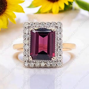 May include: A gold ring featuring a rectangular, deep red gemstone surrounded by a halo of small, clear stones. The ring's band is a simple gold design, and the setting is a mix of gold and silver. The ring is set against a soft, white background.