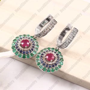 Puede incluir: Un par de pendientes de plata con un diseño circular. Cada pendiente presenta una piedra preciosa roja central, rodeada de piedras preciosas azules y verdes, y un aro de plata con pequeñas piedras transparentes.