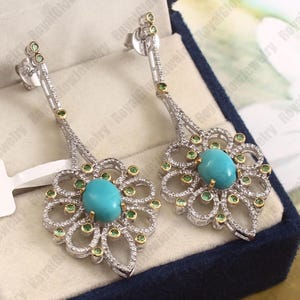 Könnte beinhalten: Silberne Ohrhänger mit ovalen türkisfarbenen Edelsteinen in einem floralen Design. Die Ohrringe haben kleine grüne und klare Edelsteine sowie goldfarbene Akzente. Ein schönes Schmuckstück.