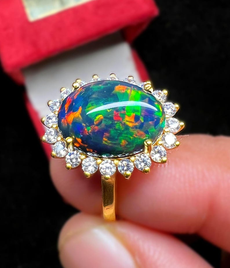 May include: A gold ring featuring a large, oval black opal gemstone with vibrant internal colors. The opal is surrounded by a halo of small, clear gemstones. The ring's band is a simple, polished gold.