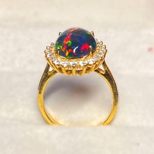 May include: A gold ring featuring a large, oval black opal gemstone with vibrant flashes of red, green, and blue. The opal is surrounded by a halo of small, clear gemstones. The ring's band is a simple, polished gold.